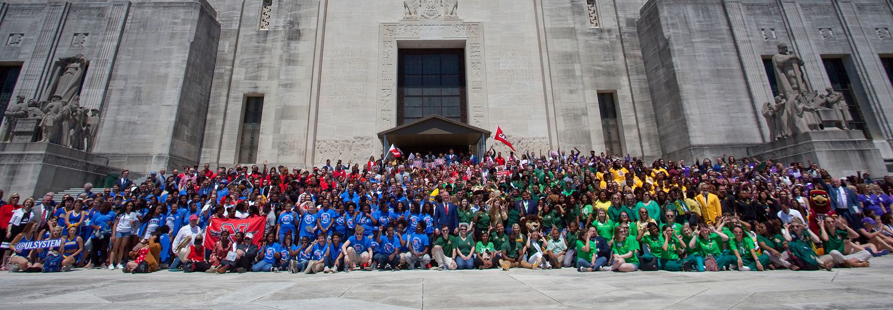 Large group of students from the nine universities that make up the University of Louisiana System, wearing school colors, gathered on the steps of the Louisiana State Capitol for a group photo.
